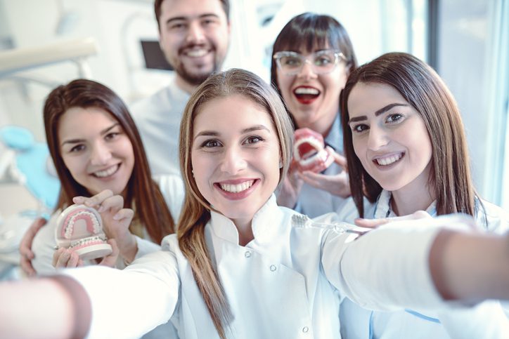 Group of cheerful dental professionals taking a selfie in a clinic, holding dental models and wearing white coats.