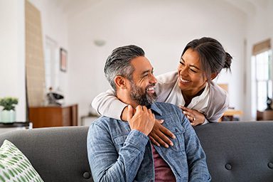 Smiling couple sitting on a couch, looking at each other warmly — highlighting the confidence that comes with a healthy smile.