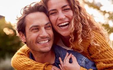 Joyful couple outdoors with woman hugging man from behind — showcasing the happiness that a bright, healthy smile brings.
